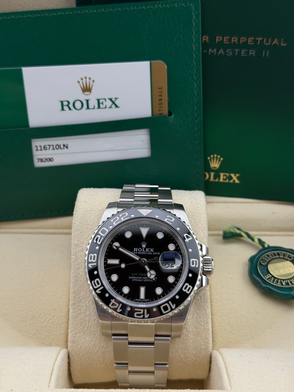 Rolex GMT-Master “Green Arrow” 116710LN - watch with black dial and silver case on a beige cushion, with Rolex branding in the background.